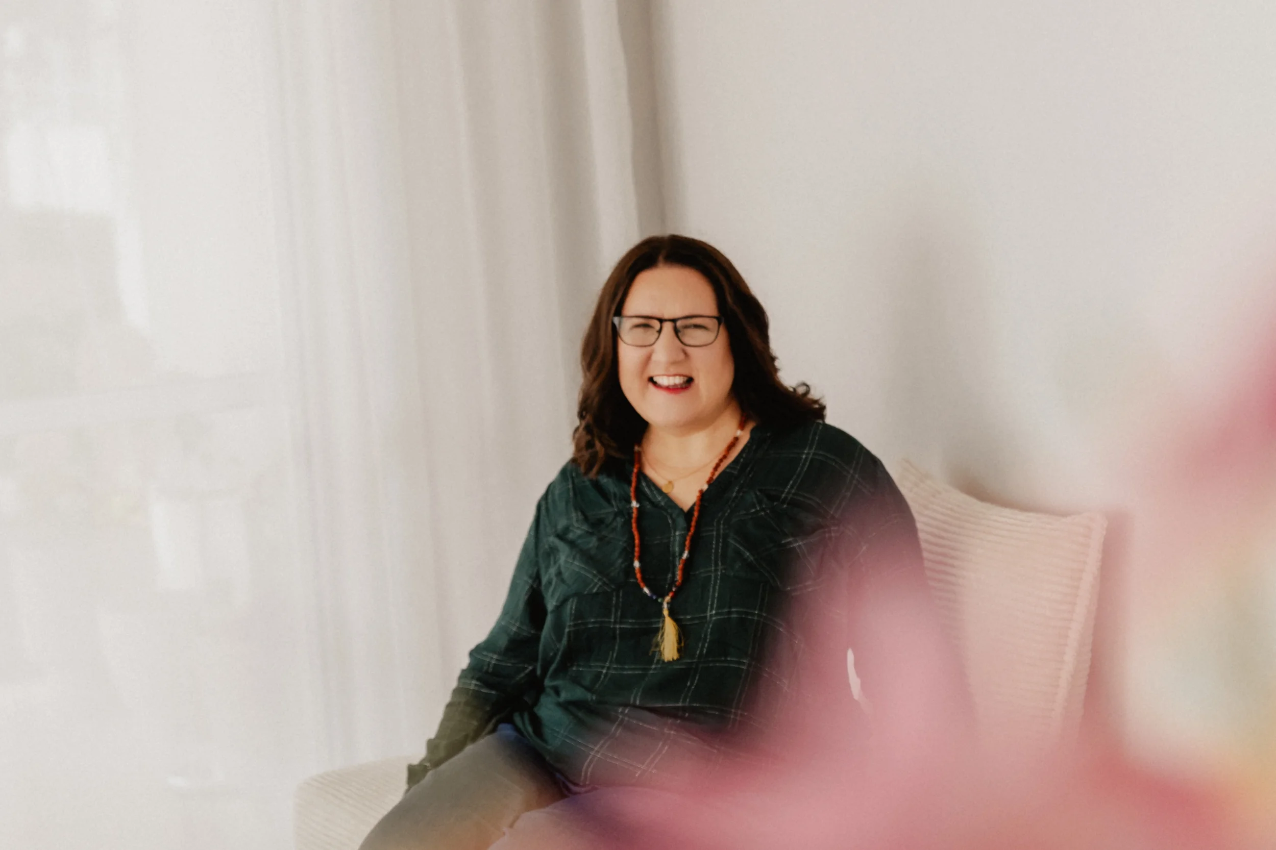 Eine Frau mit Brille sitzt auf einer gepolsterten Bank vor einem weißen Hintergrund. Sie trägt eine grüne, karierte Bluse und eine Halskette. Sie lächelt in die Kamera.