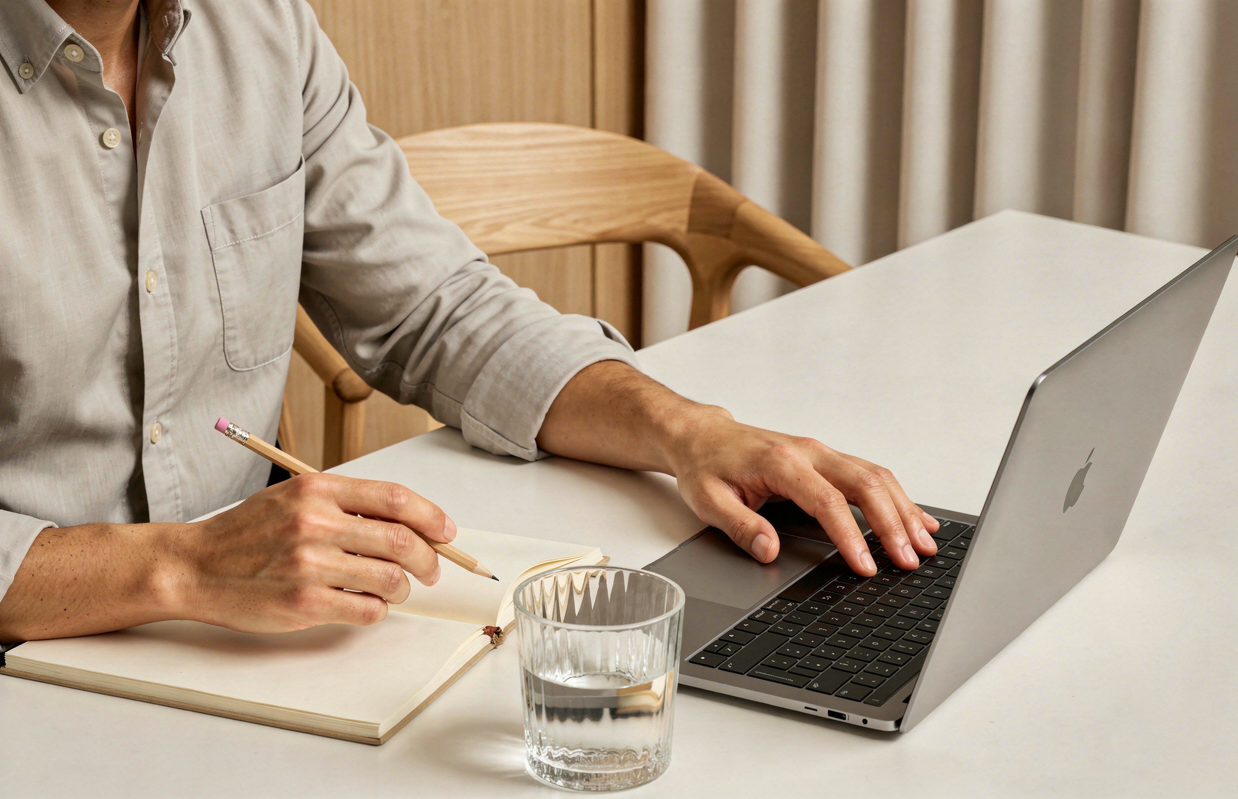 A person working at a desk with a laptop, taking notes in a notebook, and a glass of water.
