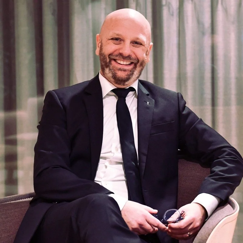 Smiling man in a black suit and tie sitting on a chair against a curtain background.