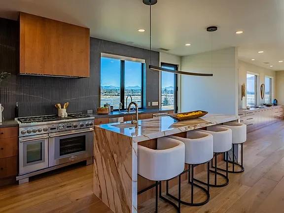 Modern kitchen with a large island, white bar stools, stainless steel stove, wooden cabinets, and large windows with an outdoor view.