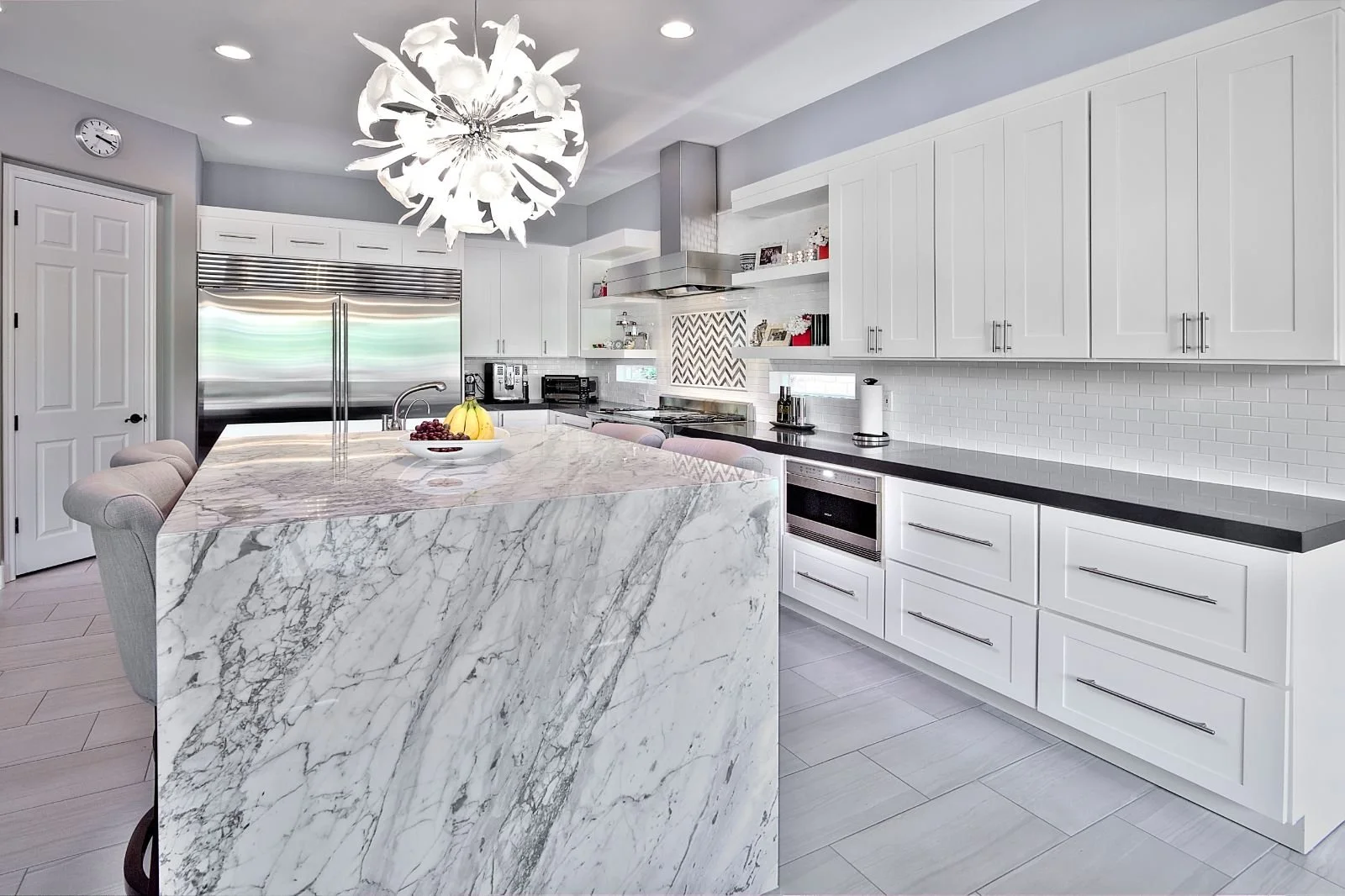 Modern kitchen with white cabinetry, black countertops, stainless steel appliances, marble island, and decorative chandelier.