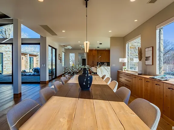 A modern dining room with a long wooden table surrounded by beige chairs, a large vase with flowers in the center, and natural light from large windows.