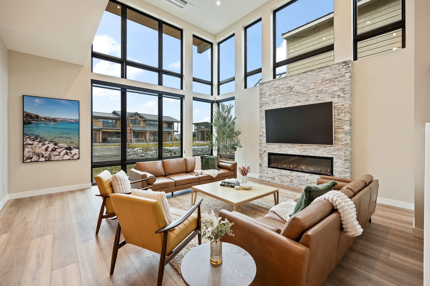 Modern living room with large floor-to-ceiling windows, tan leather sofas, a yellow armchair, a coffee table, a fireplace with a large TV above, and a view of neighborhood houses outside.