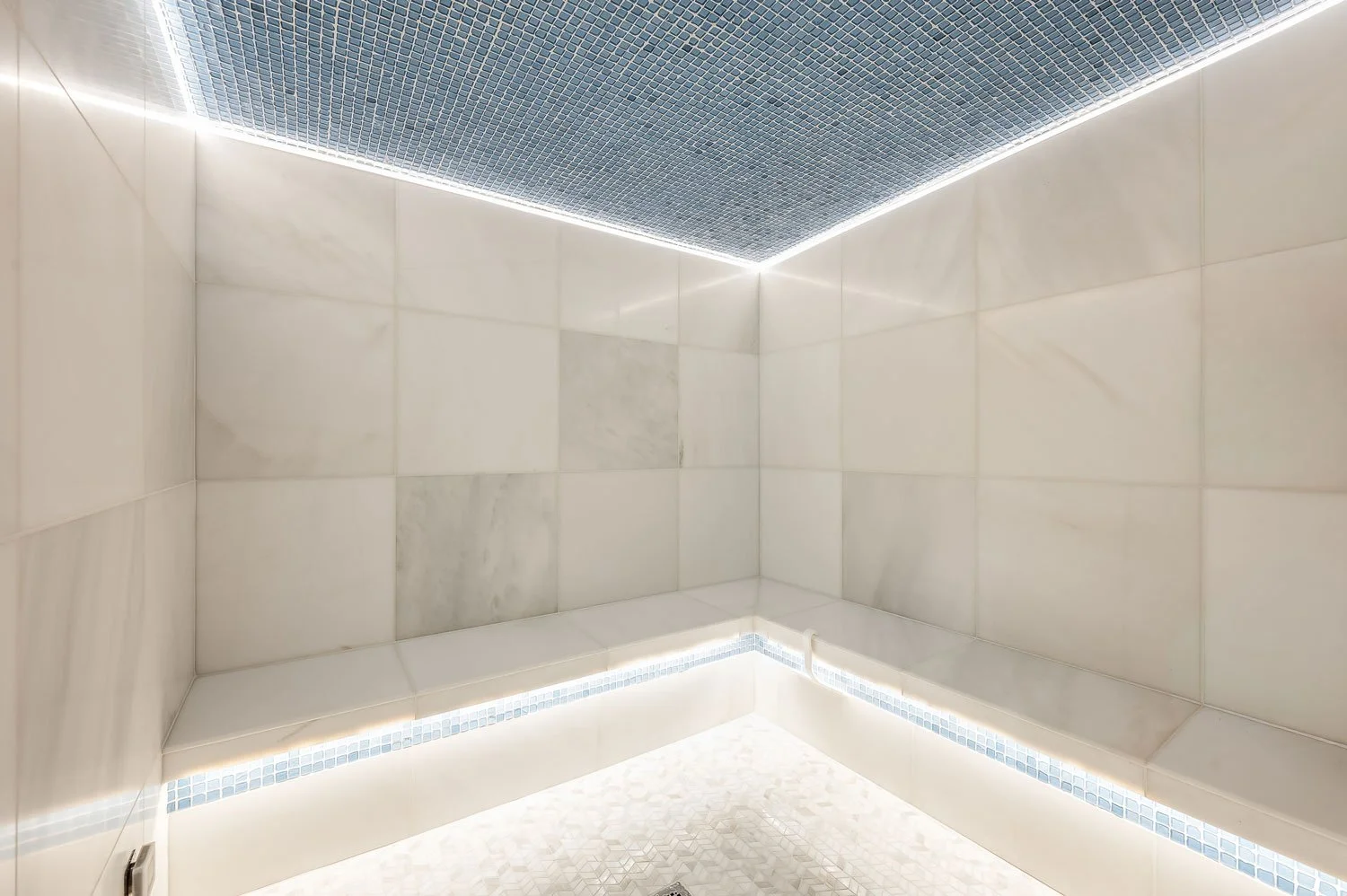 A minimalist modern spa shower with white tiled walls, blue mosaic tiles on the ceiling and along the lower edge of the walls, and soft LED strip lighting outlining the ceiling edges.