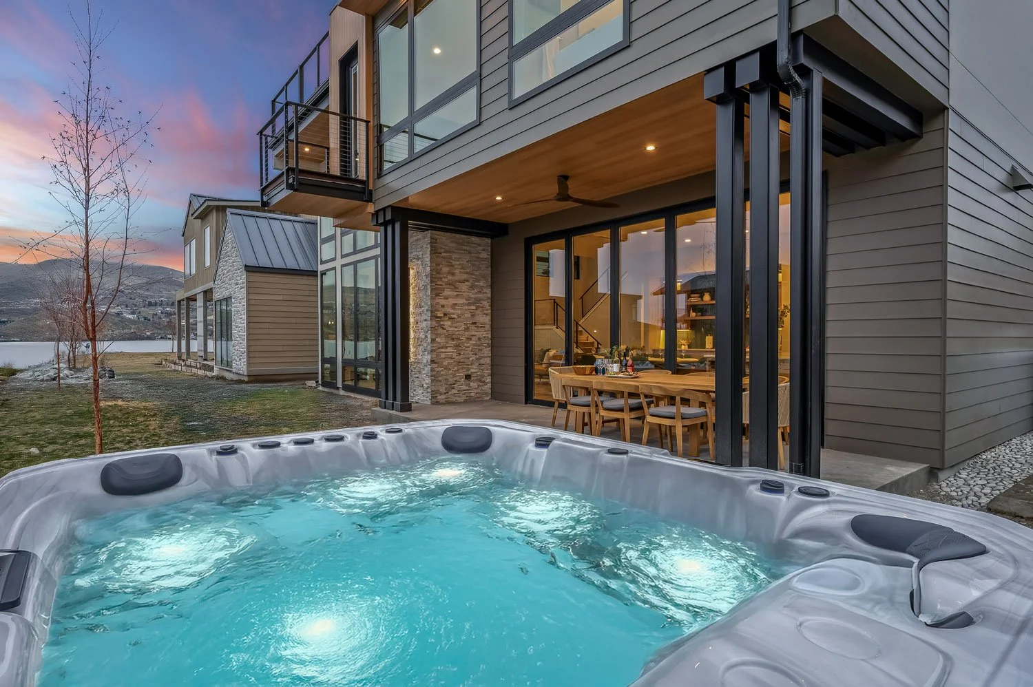Modern house with large glass windows and an outdoor deck, featuring a hot tub in the backyard, during sunset with mountain view in the background.