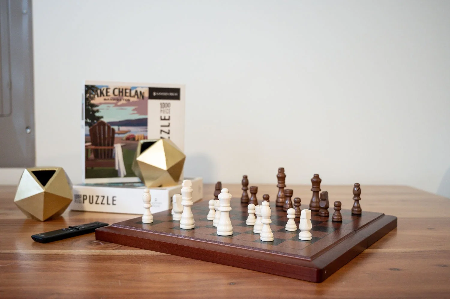 A chess game in progress on a wooden chessboard, with white and black pieces facing each other, a remote control to the left, a puzzle box behind, and decorative geometric objects on the table.