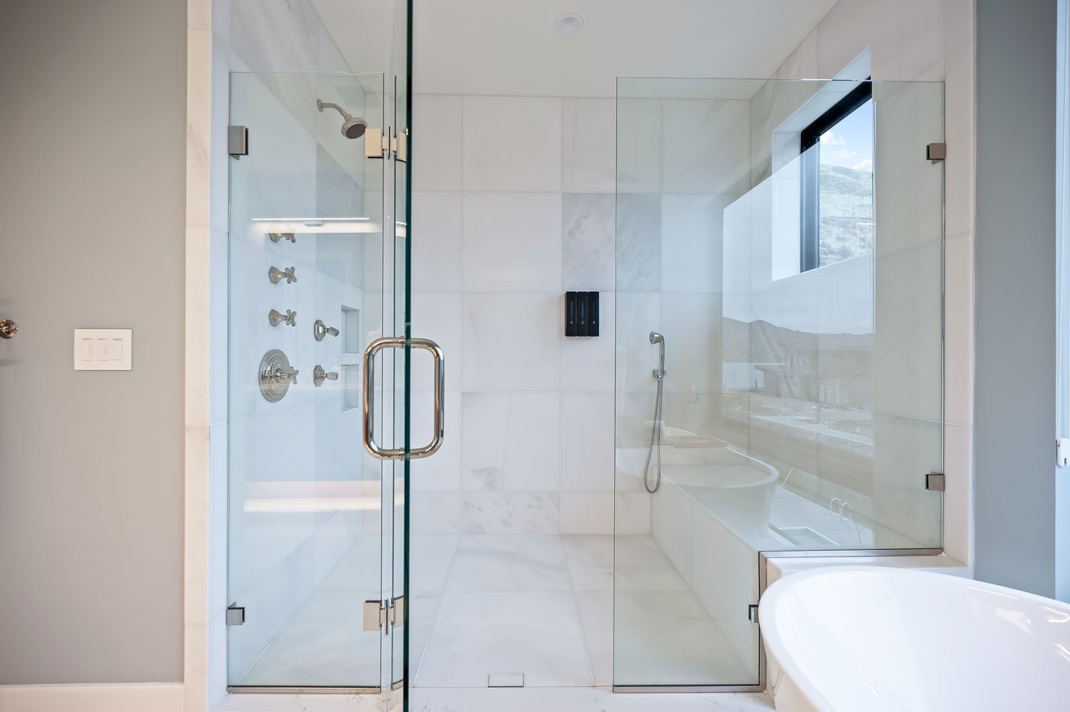 Modern walk-in shower with glass door, multiple shower controls, and a window showing outdoor scenery.