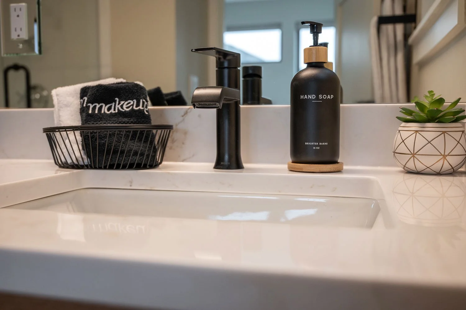 Bathroom sink with black soap dispenser, a small plant in a decorative pot, folded towels in a black basket, and a modern black faucet.