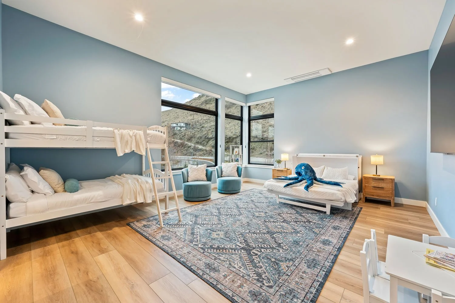 A spacious bedroom with light blue walls and hardwood floors. It features a bunk bed with white bedding on the left, a white bed with a blue octopus plush toy on top, and nightstands with lamps on either side. There's a sitting area with two round blue chairs and a patterned rug in the center, with large windows showing a mountainous landscape outside.