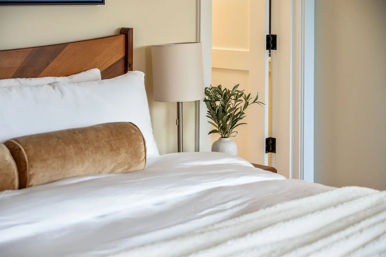 Close-up of a bedroom with a wooden headboard, white pillow, beige bolster pillow, lamp, and potted plant beside a partially open door.
