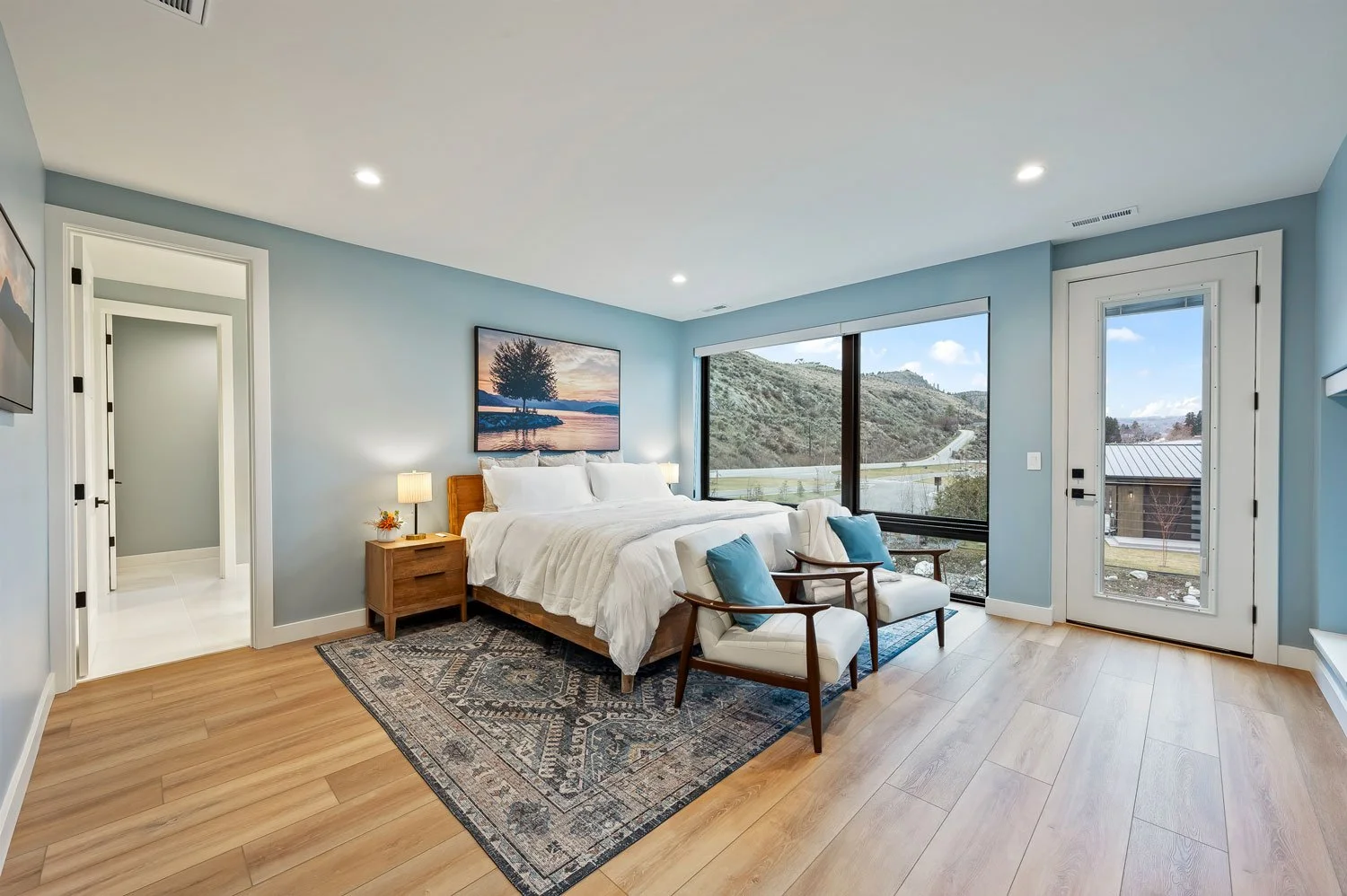 A modern bedroom with a large window showing hills and a partly cloudy sky, light blue walls, white bedding, two matching armchairs with blue pillows, a patterned rug, and a wooden nightstand with a lamp.
