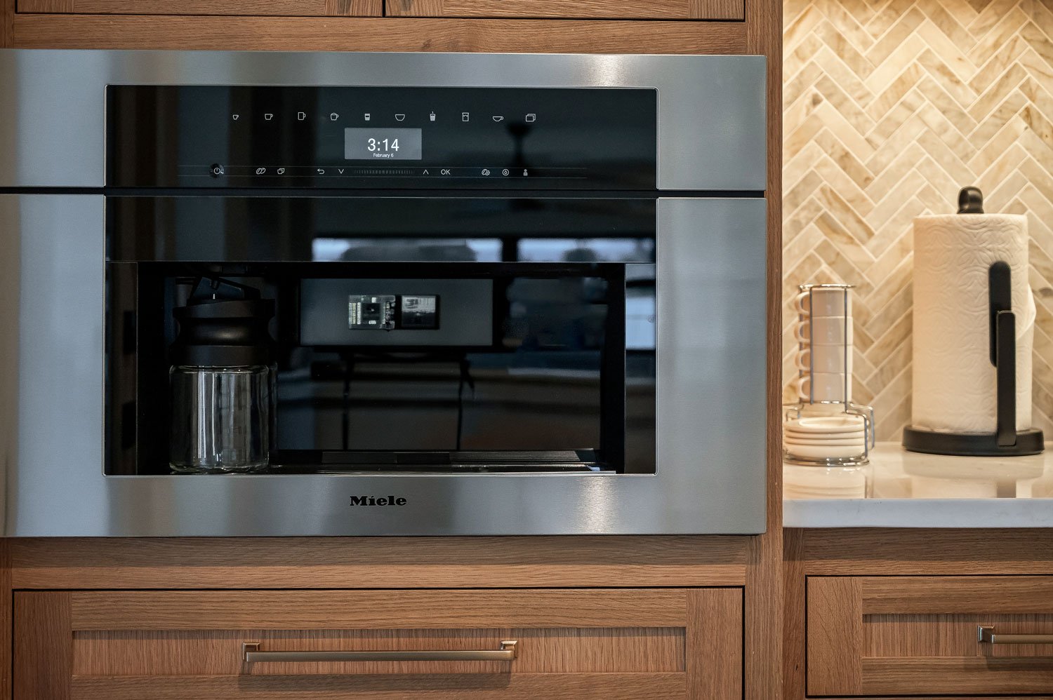 A built-in Miele coffee machine with a digital clock display, set in wooden cabinetry next to a white marble countertop with paper towel roll and containers.