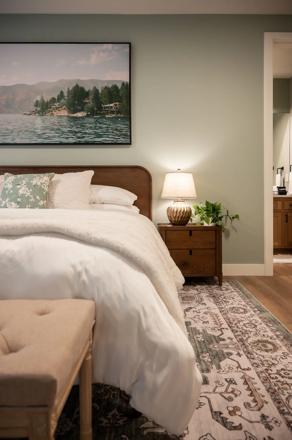 A cozy bedroom with a neatly made bed, a wooden side table with a lamp and green plant, a framed landscape photograph of a lake and mountains, and an open doorway leading to a bathroom with wooden cabinets.