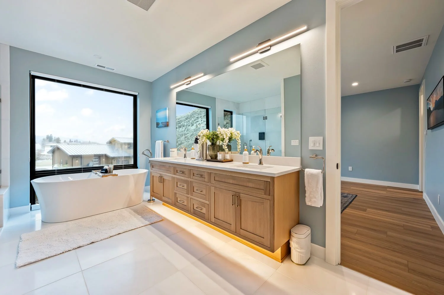 Modern bathroom with a large bathtub near a window, a wooden double vanity with a large mirror, and a glass-enclosed shower in the background.