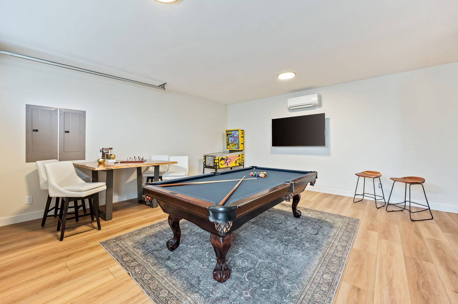 A game room with a pool table, two stools, two chairs at a table, a TV, and a pinball machine in the corner.