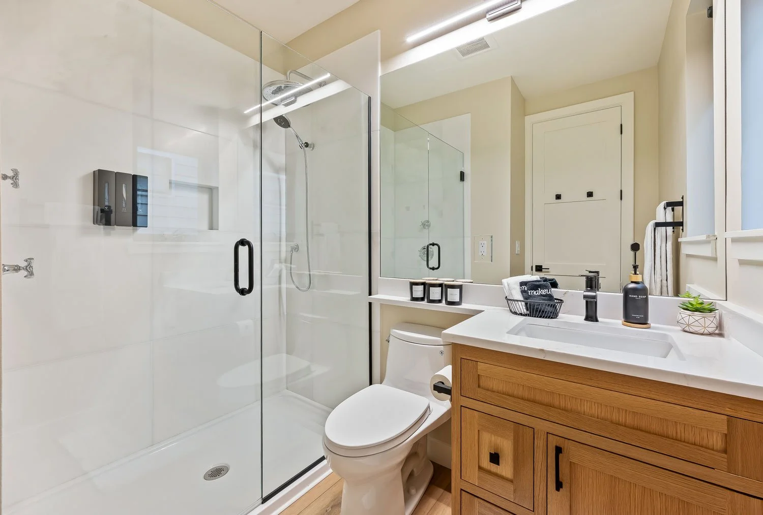 Modern bathroom with glass shower enclosure, white toilet, wooden vanity with a white countertop, black faucet, and small decorative items on the counter, including candles and a soap dispenser. There is a mirror above the vanity and a window with a white frame.