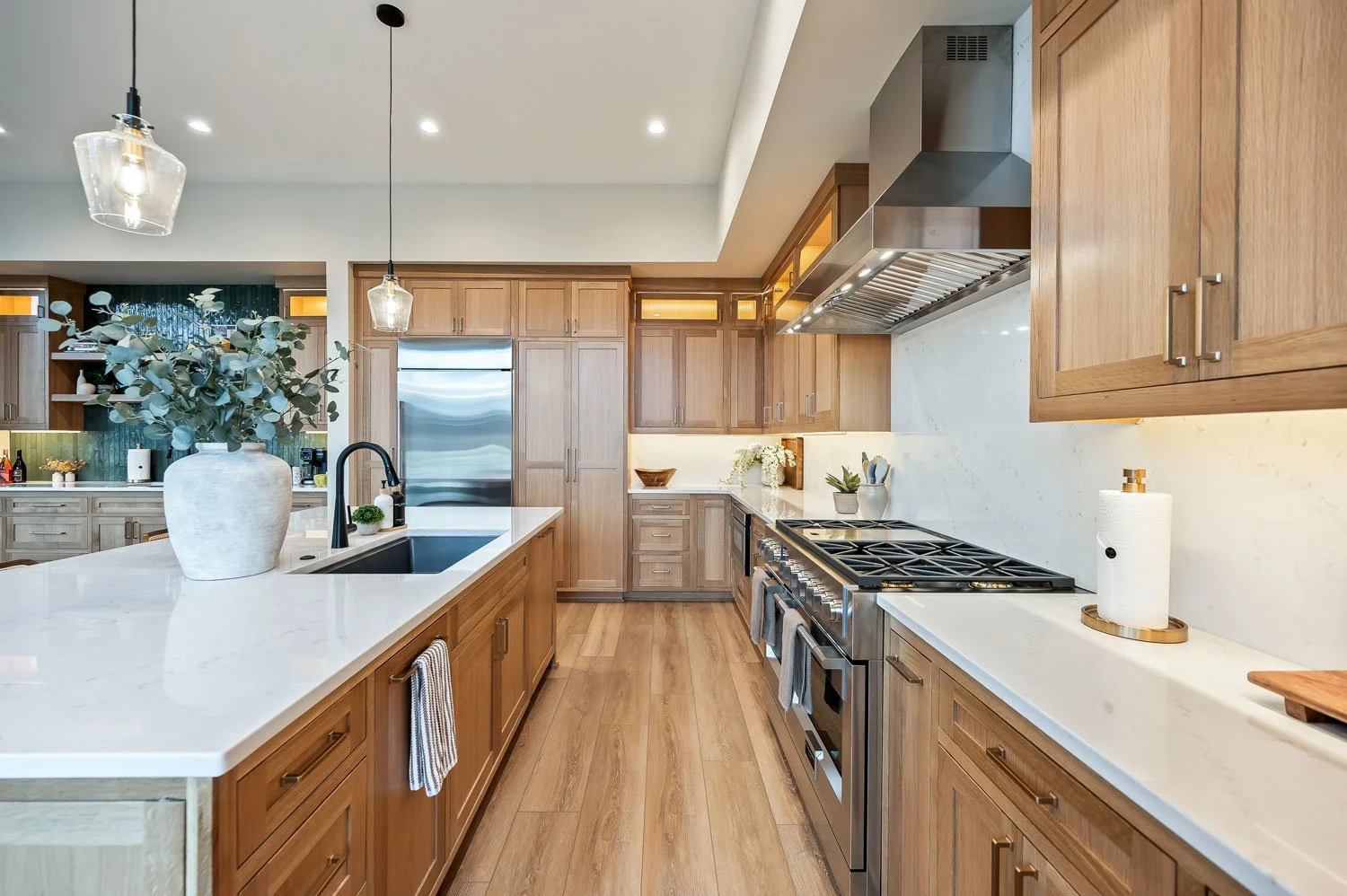 A modern kitchen with wooden cabinets, white countertops, a stainless steel stove, and a large island with a white vase and greenery.
