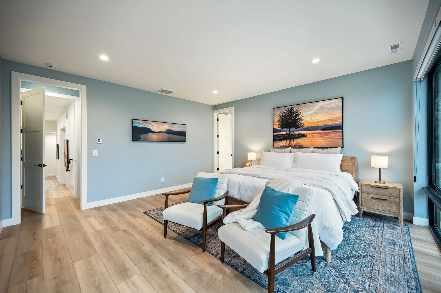 Modern bedroom with a large bed, framed landscape photos, two chairs with blue pillows, wooden nightstands with lamps, and a large window showing an outdoor scene.