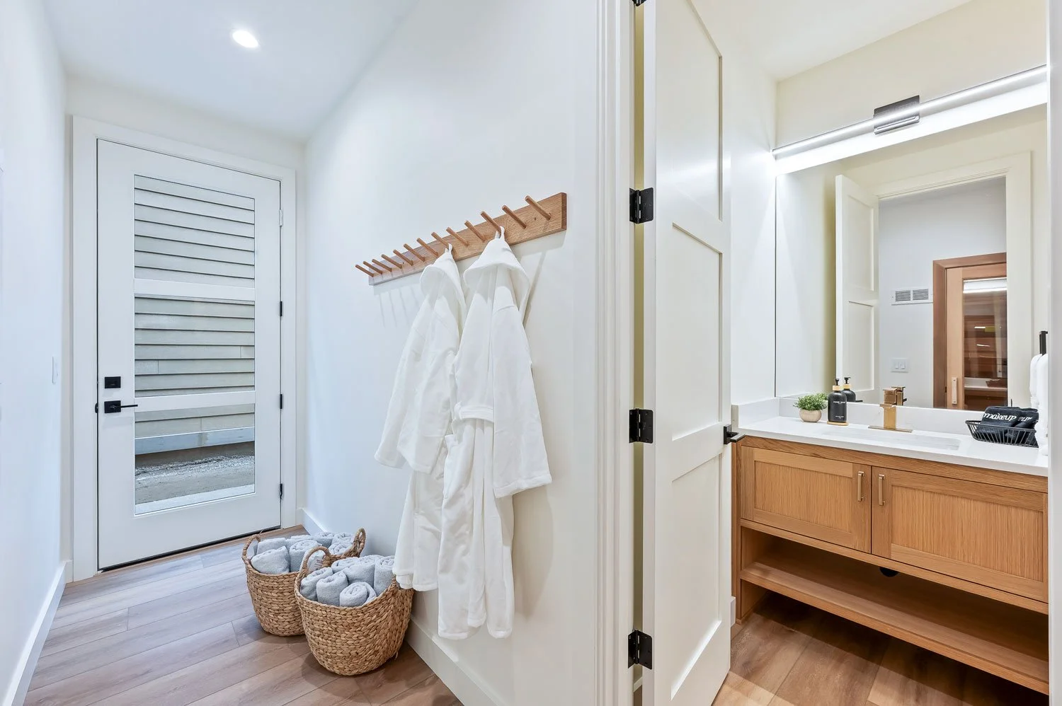 Entryway with white robes hanging on a wooden coat rack, baskets of rolled towels, and a view into a bathroom with a wooden vanity and mirror.