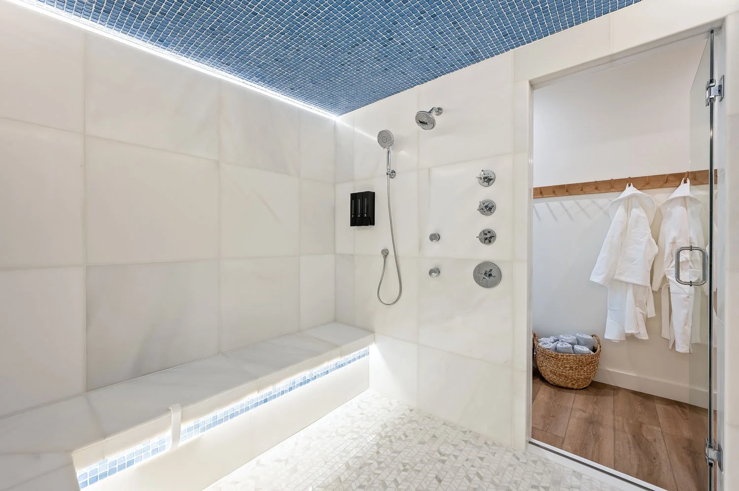 Modern walk-in shower with white tiled walls, multiple shower heads, a built-in bench, and LED lighting on the ceiling, with an adjacent changing area with robes and towels in a basket.