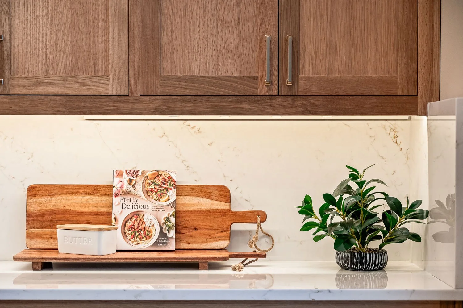 Kitchen countertop with a wooden cutting board, a book titled "Pretty Delicious," a butter dish, and a potted plant with green leaves in a black striped pot.