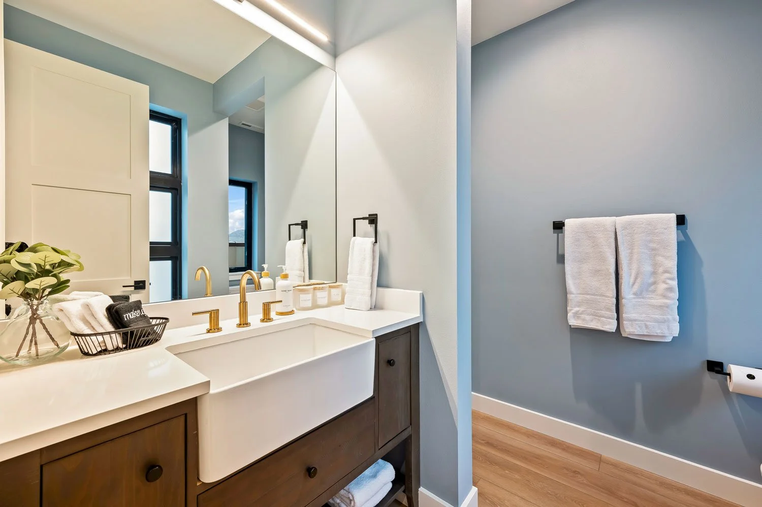 Modern bathroom with a white sink, gold fixtures, a large mirror, and light blue walls. Towel racks with white towels, a potted plant, and window with a view of mountains and sky.