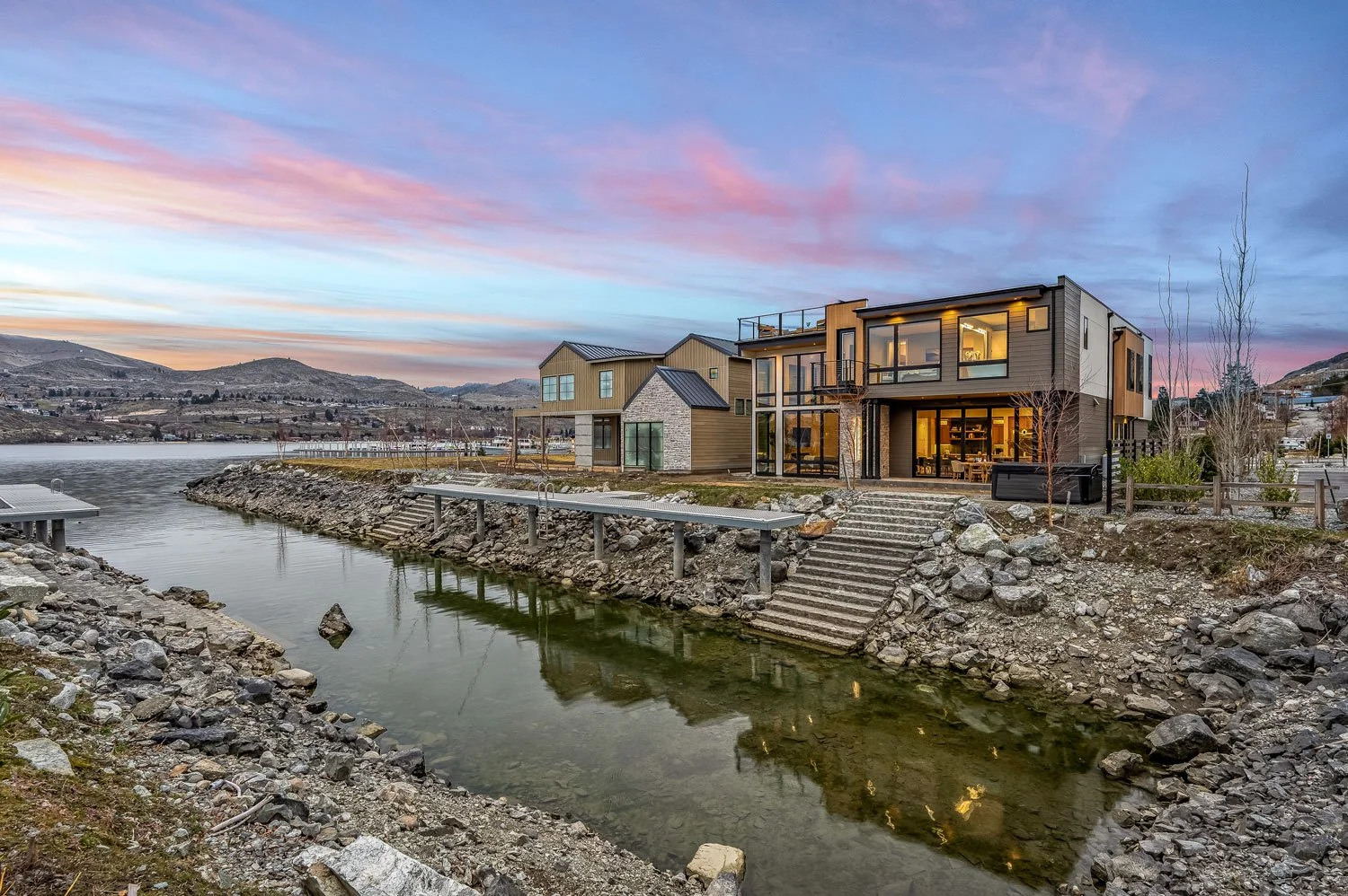Modern multi-story house with large windows by a body of water during sunset, with distant hills and colorful sky.