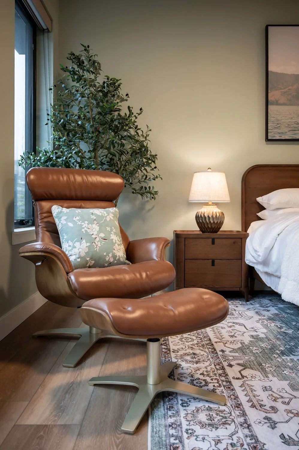 The image shows a cozy bedroom corner with a leather lounge chair and ottoman, a wooden nightstand with a lamp, a bed with white linens, a window with blinds, a large leafy potted plant, and a framed landscape picture on the wall.