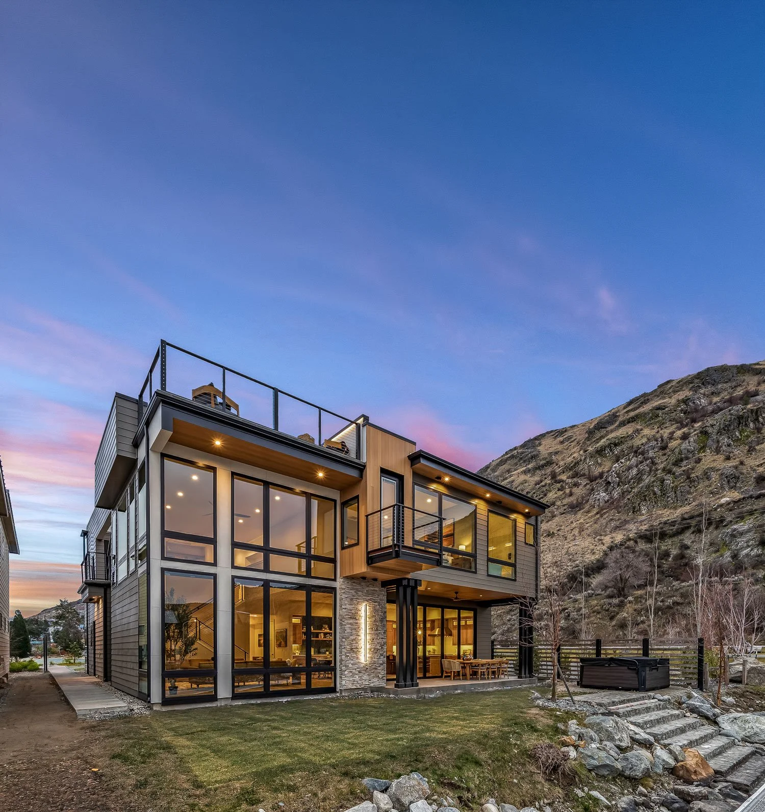 Modern two-story house with large glass windows, situated at the base of a hillside, with outdoor patio and mountain landscape in the background during sunset.