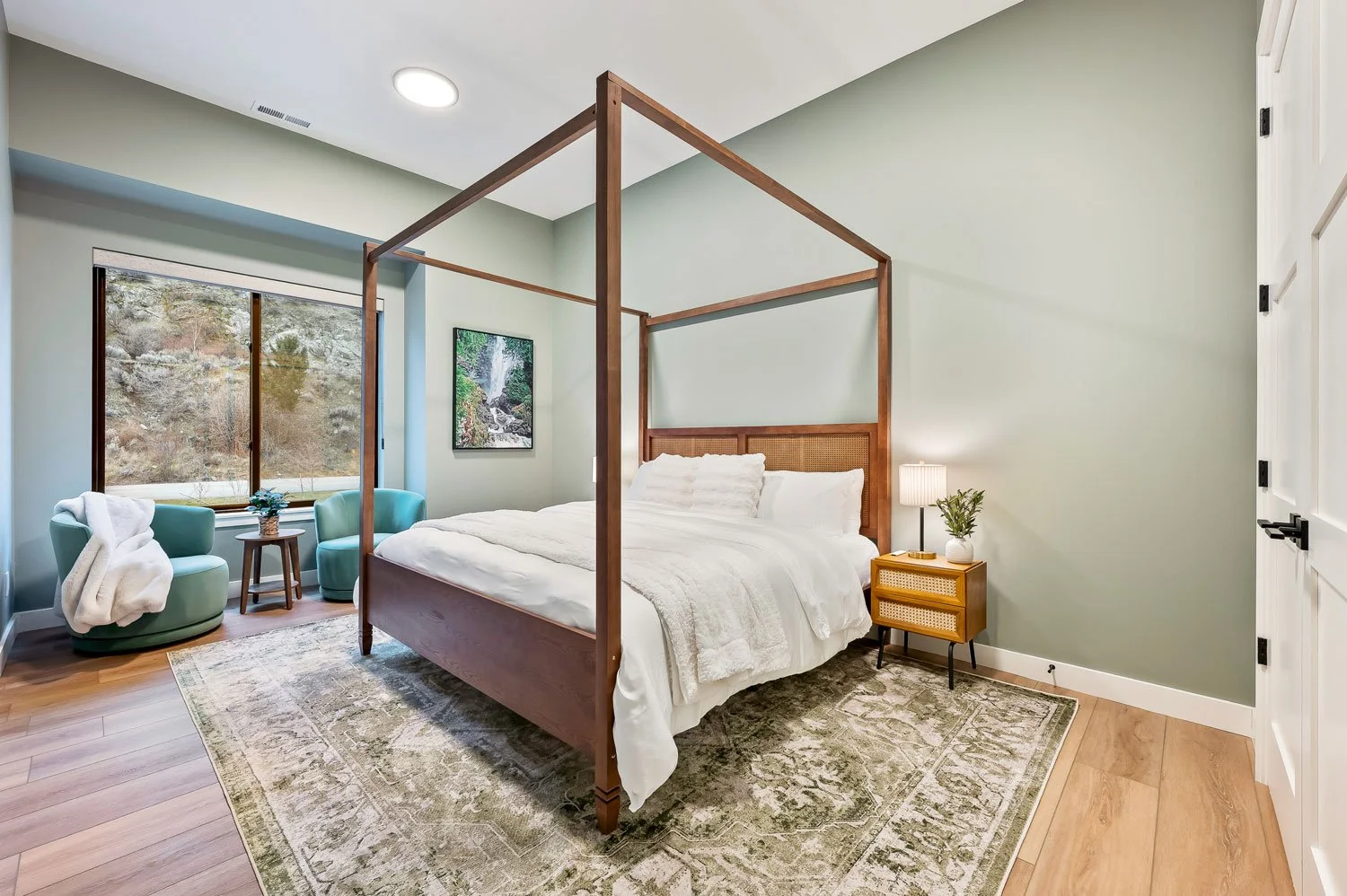 Bedroom with a four-poster wooden bed, nightstand with lamp and plant, two green armchairs by a large window with outdoor view, framed waterfall picture on wall, light green walls, hardwood floor, and patterned area rug.