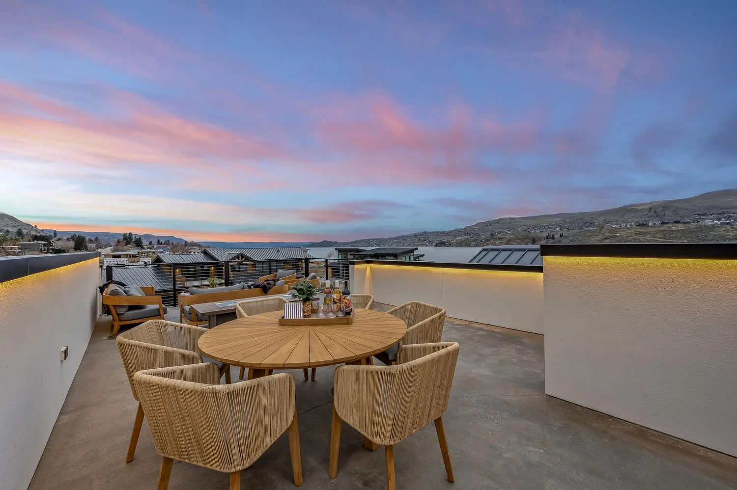 Rooftop patio with a round table and chairs, overlooking a scenic landscape with hills and a colorful sunset sky.