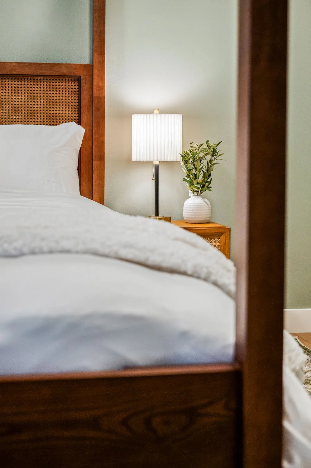 A bedroom scene featuring a bed with a white comforter, a wooden headboard, a bedside table with a lamp, and a potted plant.