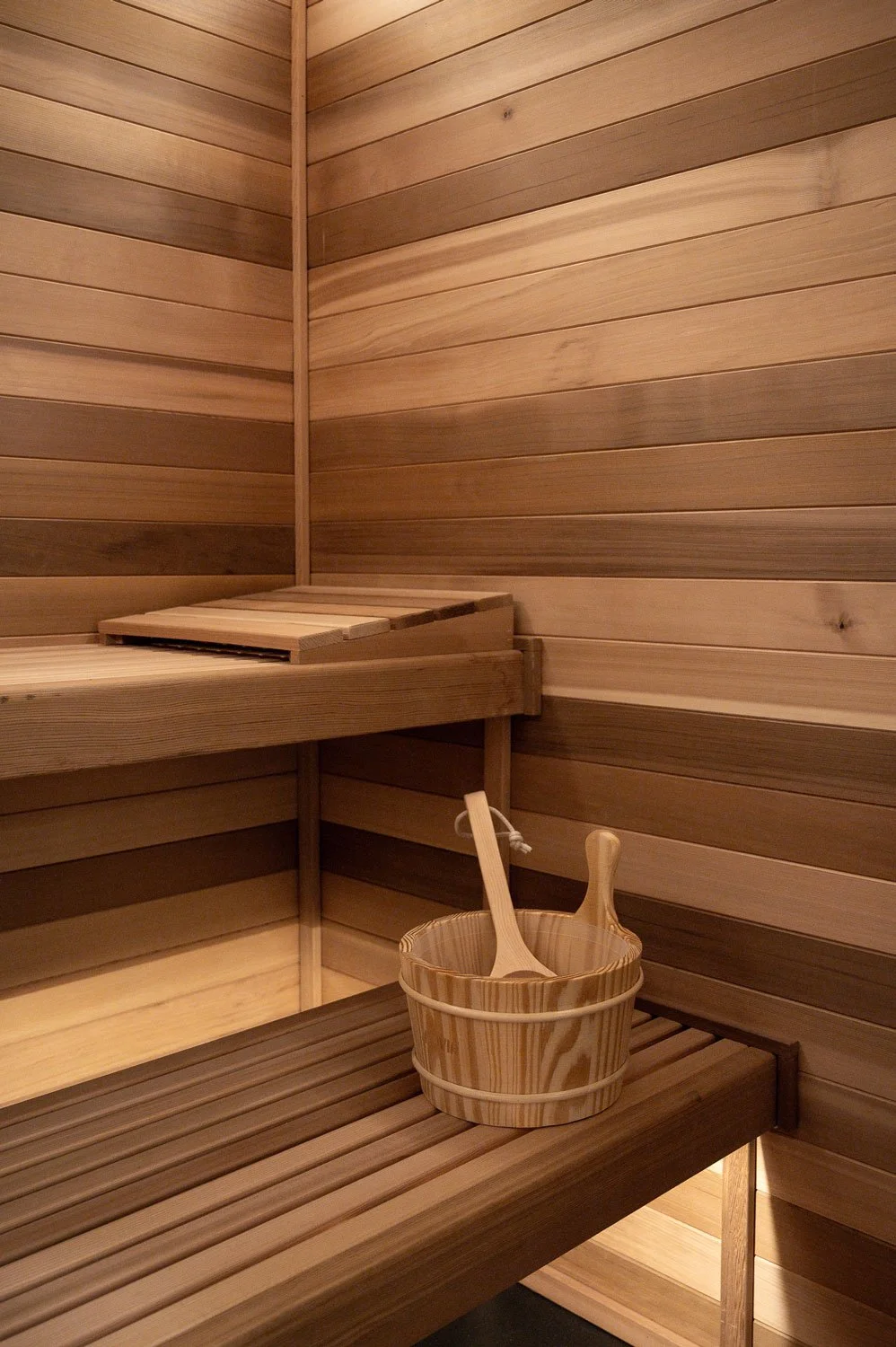 An interior view of a wooden sauna with horizontal wooden planks on the walls and benches. A small wooden bucket with two wooden utensils inside is on the lower bench, and a wooden step is on the upper bench.
