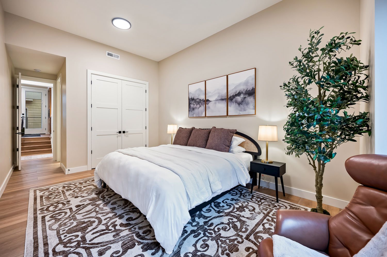 A modern bedroom with a large bed, white bedding, and three brown pillows. There are two lamps on black nightstands on either side of the bed. A triptych of watercolor landscape art hangs above the bed. To the right, there's a large green potted plant, and in the bottom right, part of a brown leather chair. The room has wood flooring and a patterned area rug.