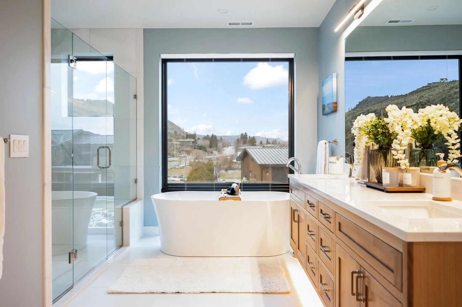 Modern bathroom with a large window showing a mountain view, featuring a freestanding bathtub, a glass-enclosed shower, wooden vanity with a white countertop, and decorative flowers.