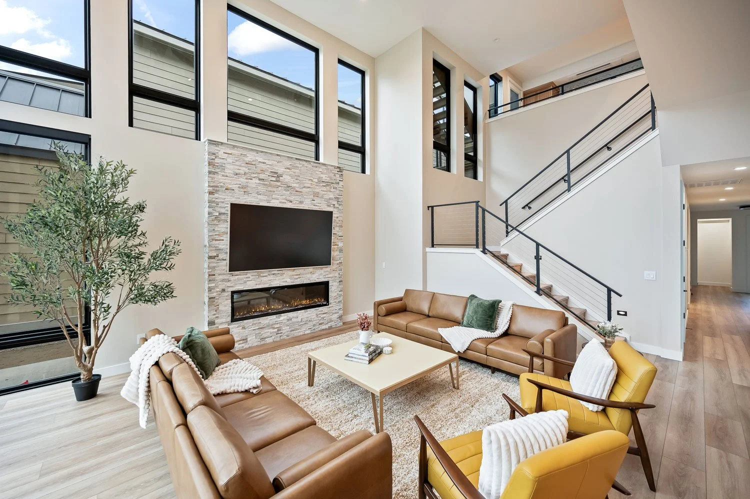 Modern living room with large windows, a stone fireplace with a mounted flat-screen TV, beige sofas, yellow armchairs, a coffee table, a potted plant, and a staircase with black railings.