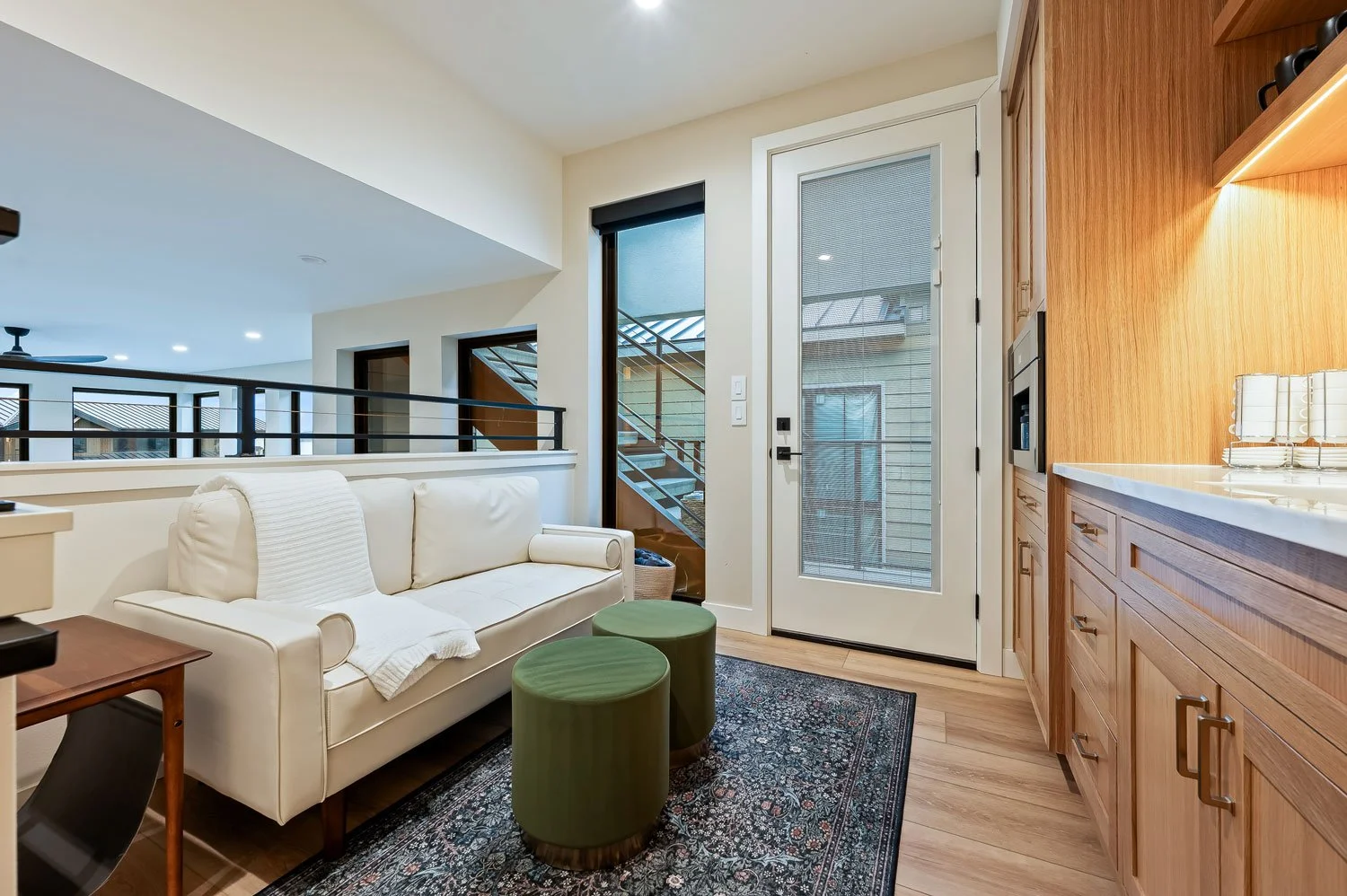 Living room view with a white sofa, green ottomans, a wooden side table, hardwood floors, a patterned rug, a glass door to a patio, and wooden kitchen cabinets.