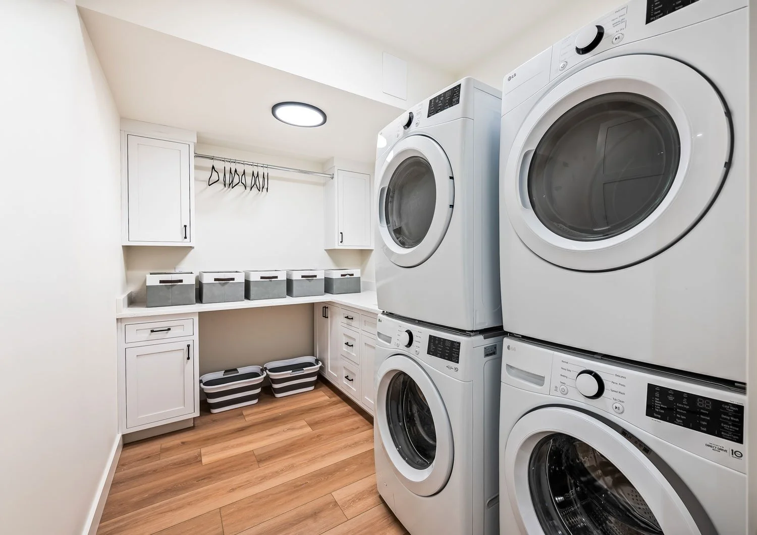 A laundry room with four white front-loading washing machines stacked on two stacks, white cabinets, storage boxes, and a clothing rod with hangers, and a wooden floor.