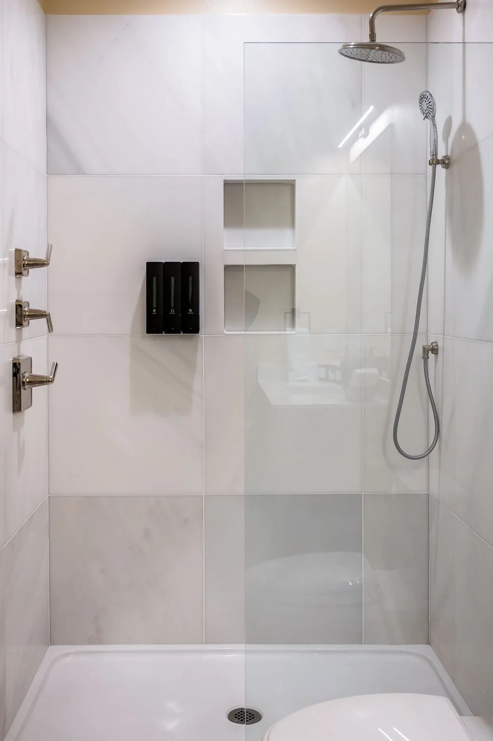 A modern shower area with white tiled walls, a glass door, a rainfall showerhead, a handheld showerhead on a hose, built-in niche shelves, and three black soap dispensers on the wall.