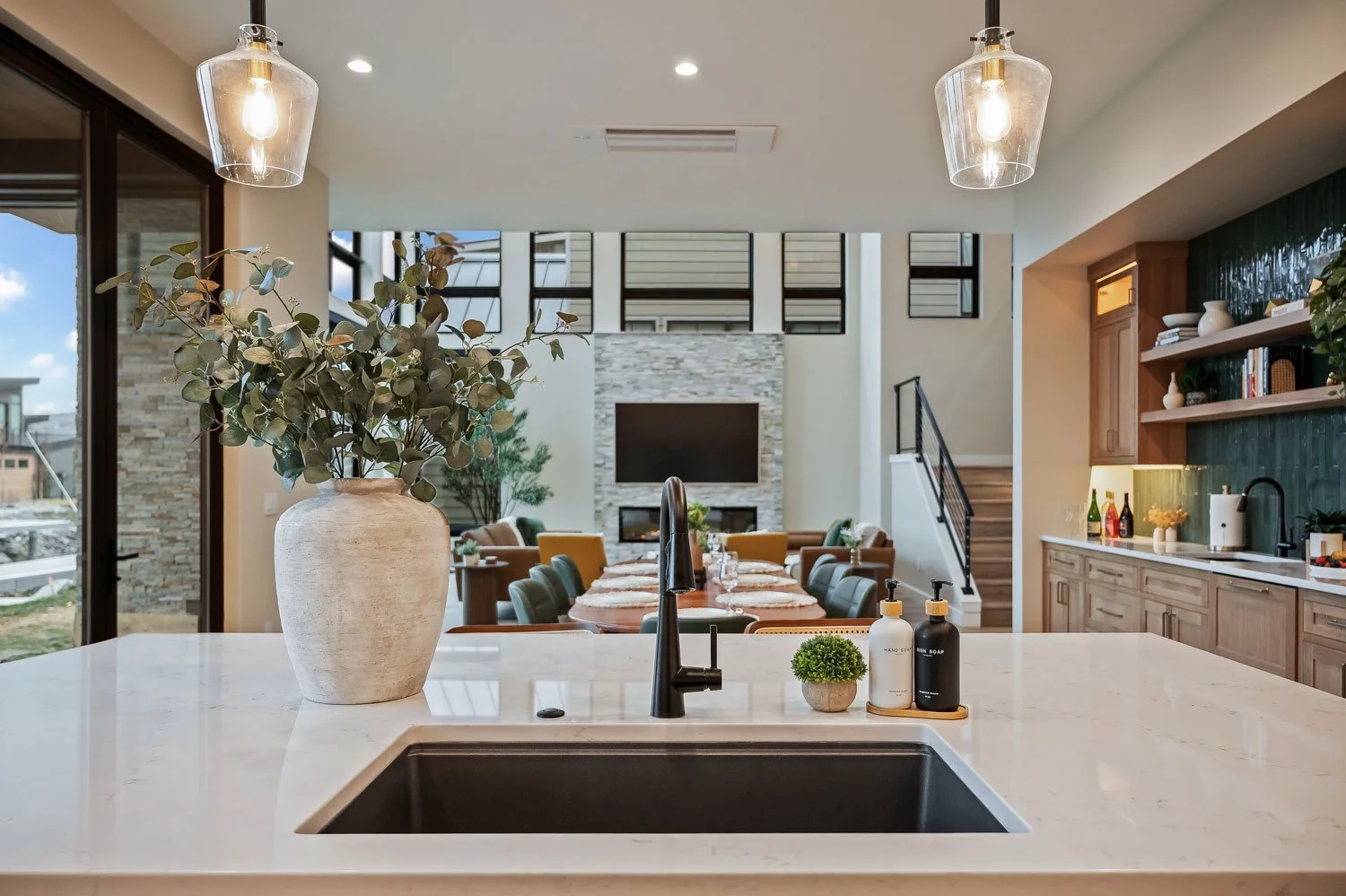 Modern kitchen with a white marble countertop, large white vase with green foliage, black sink, and soap bottles. Open living area with dining table, chairs, and a TV mounted on a stone wall.