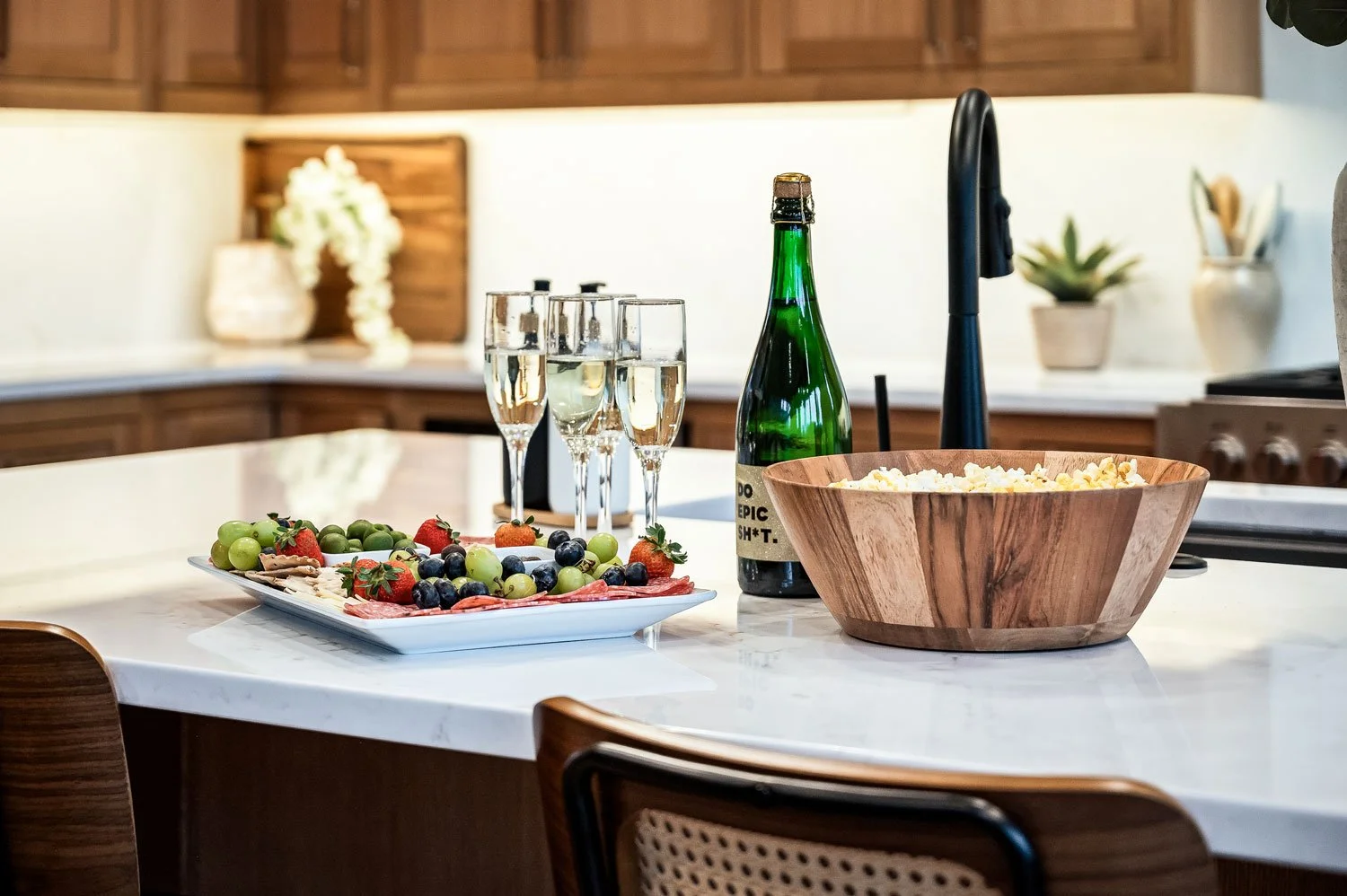 A kitchen countertop with a wooden bowl of popcorn, a bottle of sparkling wine, five glasses of champagne, a plate of mixed grapes, strawberries, blueberries, and slices of prosciutto, with a black faucet and decorative plants in the background.
