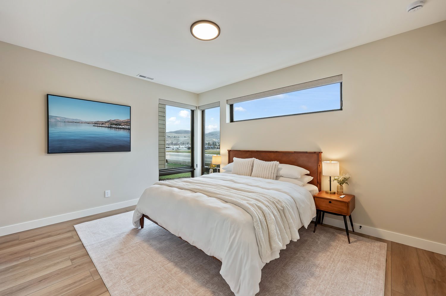 Modern bedroom with a large bed, white bedding, wooden headboard, nightstand with a lamp and flower, window with a city view, and wall-mounted TV.