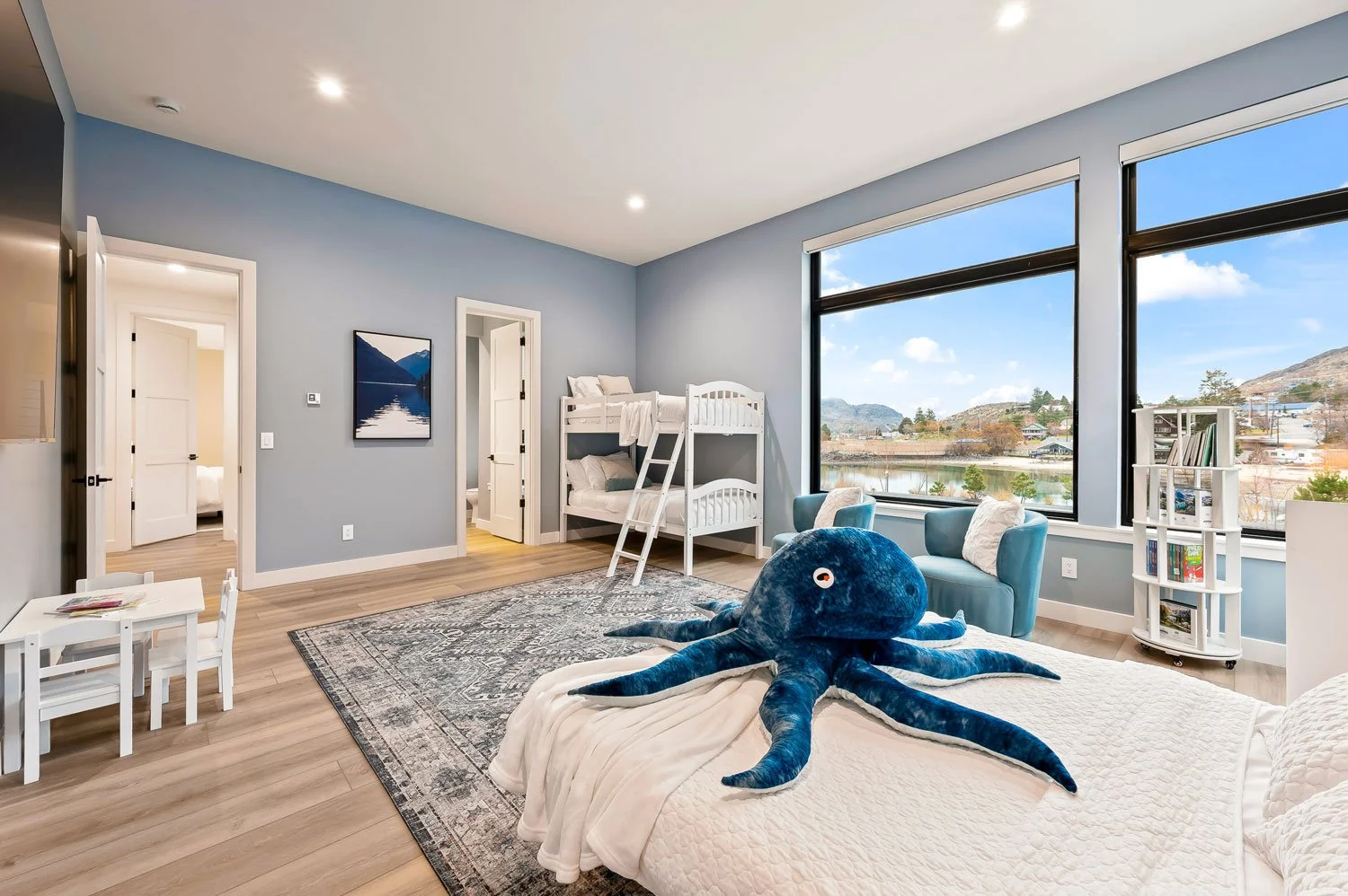 Children's bedroom with bunk beds, large windows showing outdoor scenery, a stuffed octopus on the bed, blue armchairs, a small white table and chairs, a bookshelf, and artwork on the wall.