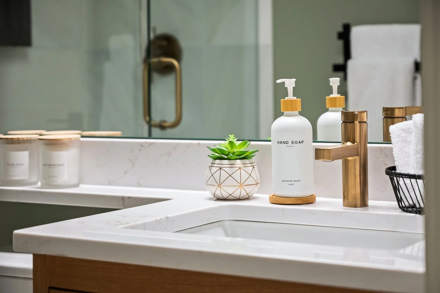 Bathroom sink countertop with hand soap, a small potted plant, a folded towel, and toiletries, reflected in a mirror with a glass shower door in the background.