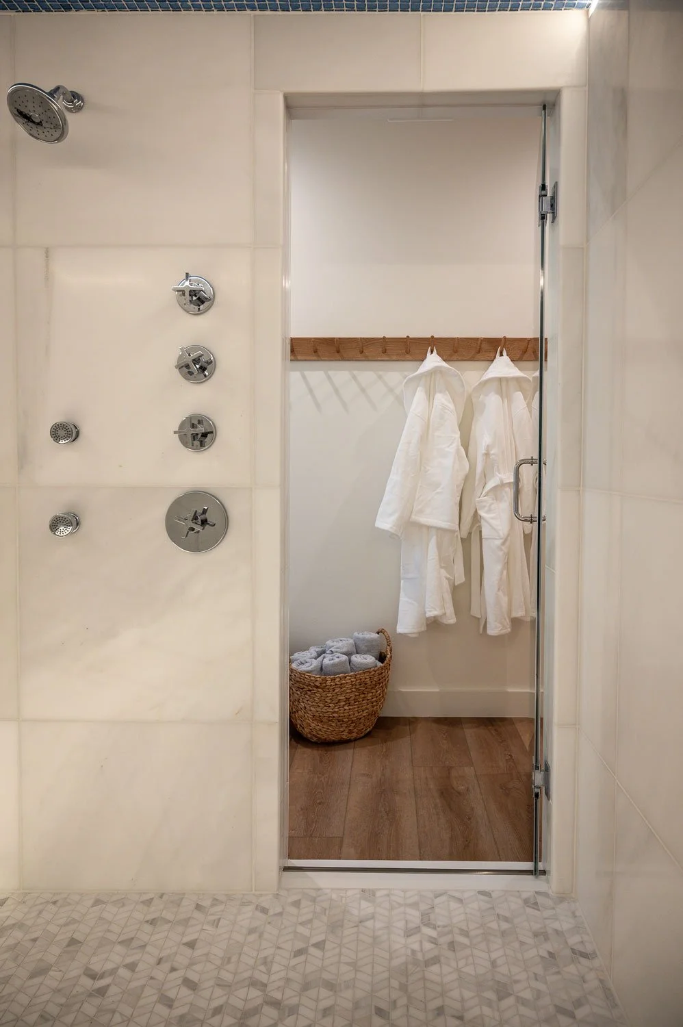 View of a shower area with multiple showerheads on the left wall, and a door opening into a space with white robes hanging on a wooden rack and a basket of rolled towels on the floor.