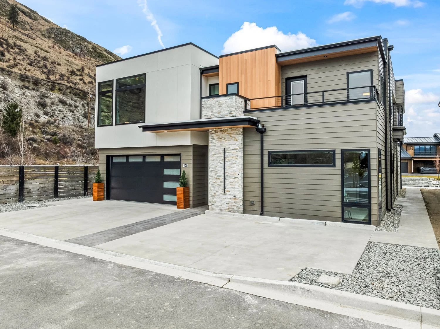 Modern multi-story house with a concrete driveway, black garage door, and a mix of beige, white, and wood exterior panels, set against a hillside with a partly cloudy sky.