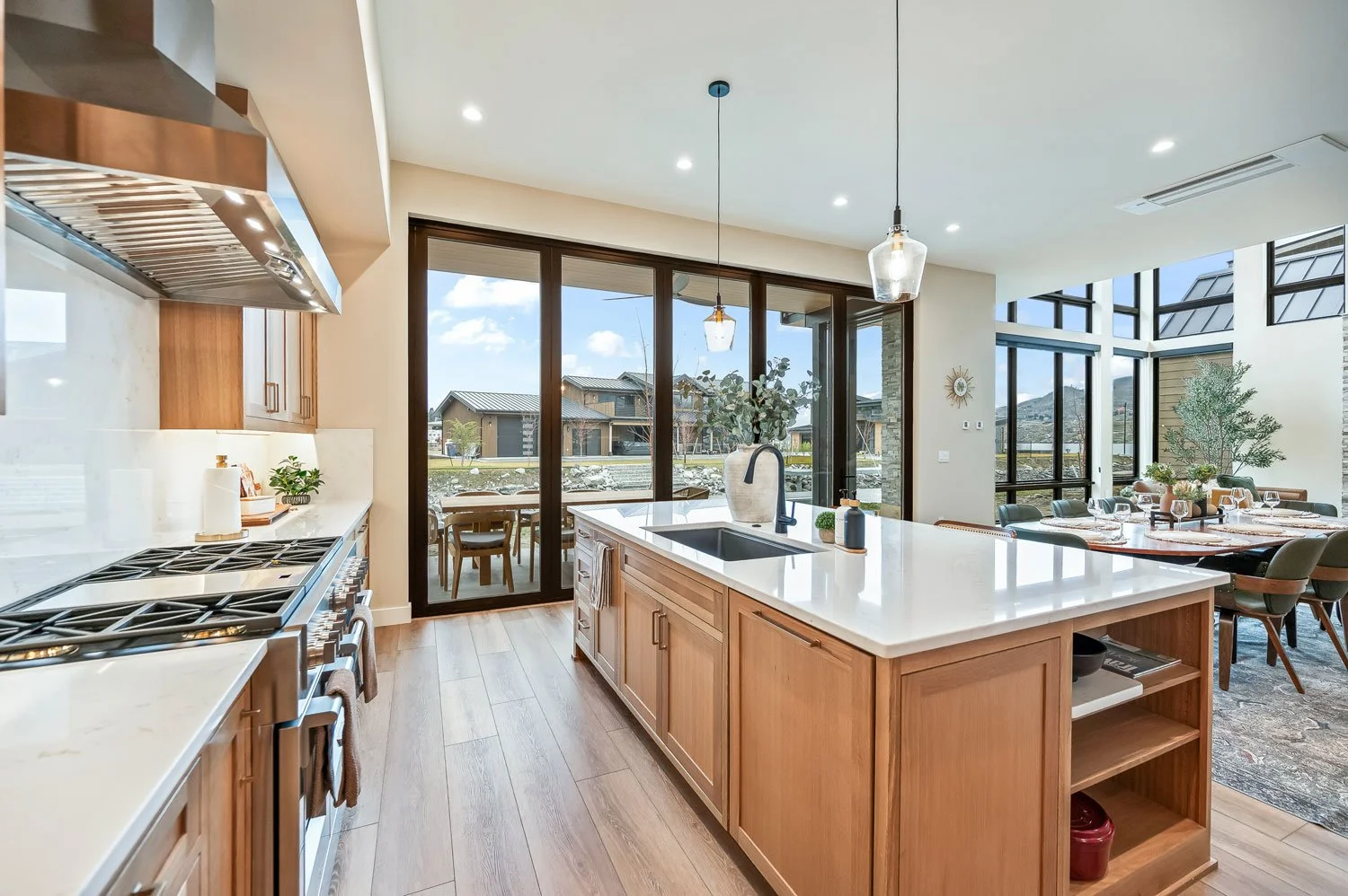 Modern kitchen with wooden cabinets and a large white island, stainless steel stove, and large windows revealing an outdoor patio and neighboring houses.