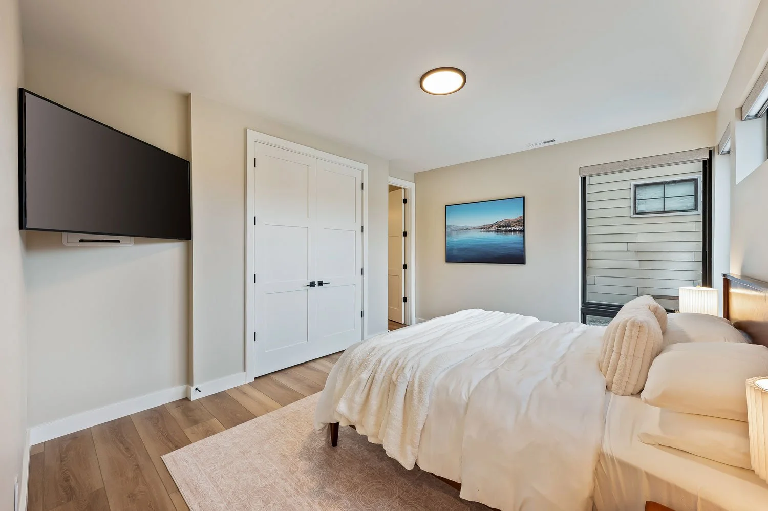 A bedroom with a bed, a wall-mounted TV on the left, a framed picture of a coast and water on the right wall, and a window with blinds. The room has beige walls, hardwood floors, and simple decor.