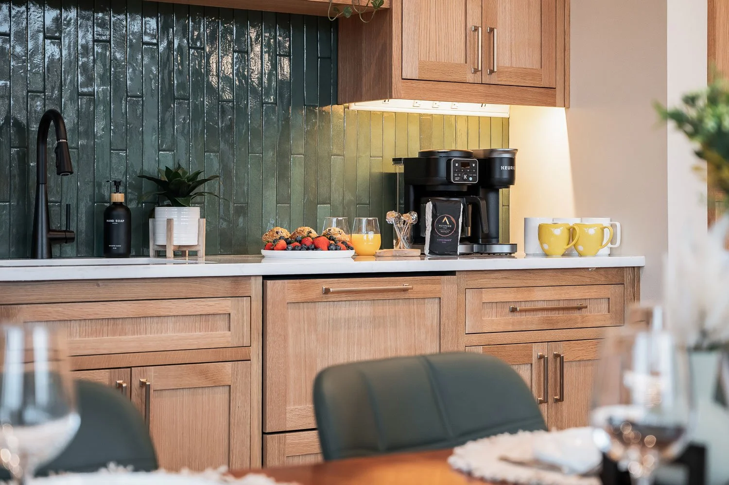 Kitchen countertop with coffee maker, glass of orange juice, bowl of mixed berries, yellow mugs, soap dispenser, and plants, with wooden cabinets and green tiled backsplash.