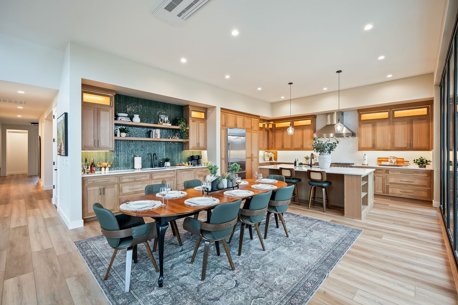 Open-concept kitchen and dining area with wooden cabinets, a central island, a stainless steel range hood, and a wooden dining table set for a meal with eight chairs, decorated with plates, glasses, and flowers.